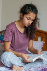An autistic teenage girl sitting on her bed reading a book.