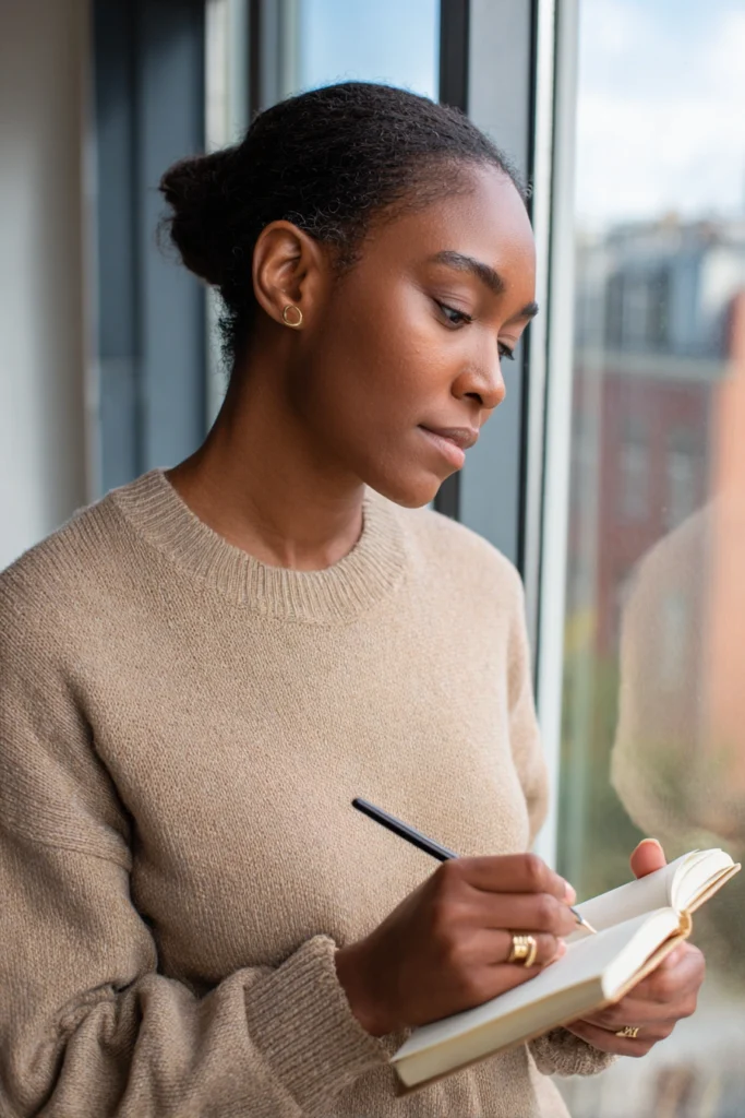A serious, slightly anxious woman writing in a diary while she stands by a window in her apartment.
