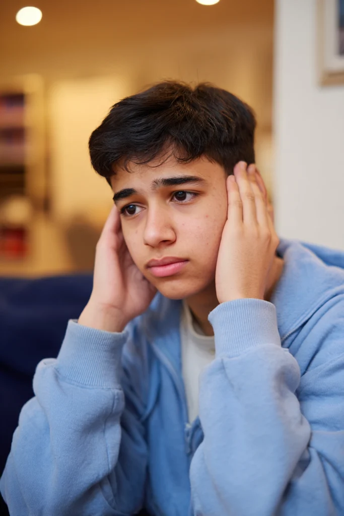 A fourteen-year-old boy in his living room, covering his ears with his hands.